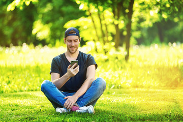 Young hipster man is sitting with cellphone at green park background and smiling.