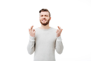 Young man making a wish emotionally with finger crossed gesture over white background. Guy praying with eyes closed isolated