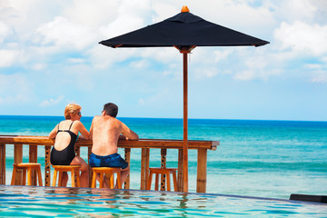 Successful retirement recreation, summer vacation concept. Retired mature couple enjoying beautiful sunny day in swimming pool at beach club. Happy senior woman and man sitting at poolside beach bar.