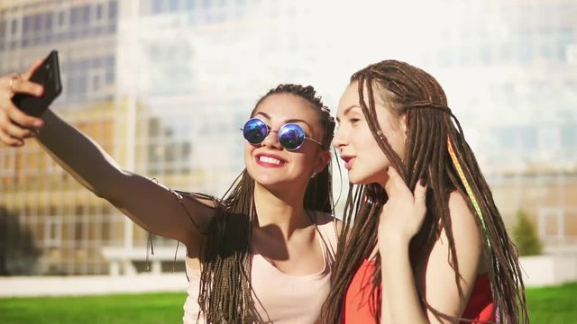 Happy women with dreads sitting on grass in summer park and talking selfies. Young friends talking and taking photos, posing outdoors and having fun. Slowmotion shot