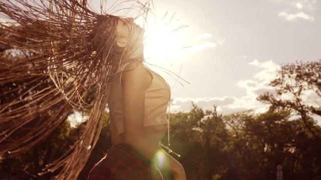 Beautiful woman with dreads styling hair and shaking her head in the sun. Happy calm attractive girl with long dreads having fun in the park, enjoying nature