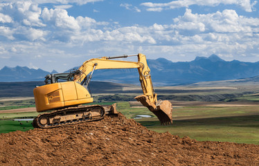 horizontal image of a yellow backhoe working on a big pile of dirt with the scoop open with beautiful blue mountains in the background in the summer time.