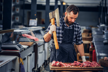 Bearded butcher cuts meat with axe.