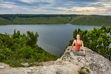 Beautiful woman is practicing yoga on the rock above big river against sky.
