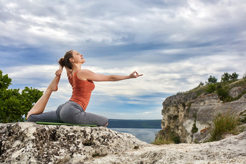 Young woman doing yoga exercise pigeon asana on the rock against river.