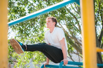 Fototapeta premium Strong young athlete. Strong young athlete is engaged in on an abandoned sports field. Athletic man. Exercises on uneven bars. Athlete Outdoors. Toned image.