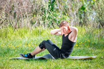 Attractive fit young man stretching before exercise, sunrise early morning backlit. Sit ups fitness man exercising sit up outside in grass in summer. Male athlete working out cross training in summer