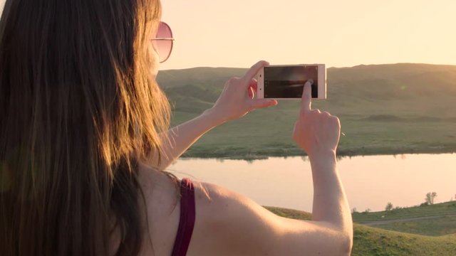 Close up from the back of girl or woman taking photo of sunset lake view landscape river at the background with her iphone. Almaty.