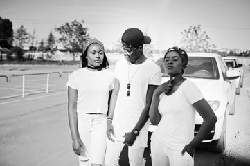 Three stylish african american friends, wear on white clothes against two luxury cars. Street...