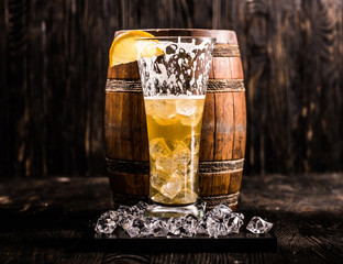 half empty glass of beer with lemon and ice with little beer barrel on the background