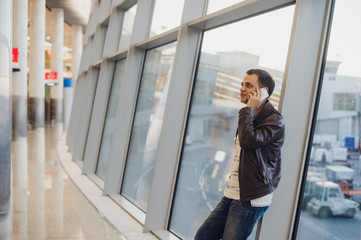 Portrait of young handsome person wearing casual style clothes standing near window in modern airport terminal. Traveler making call using smartphone.