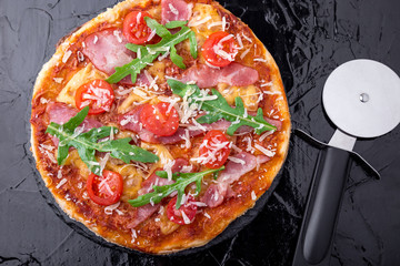 Homemade pizza with prosciutto, tomato, arugula on black slate board. Top view.