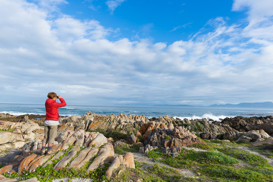 Tourist looking with binocular on the rocky coast line at De Kelders, South Africa, famous for whale watching. Winter season, cloudy and dramatic sky.