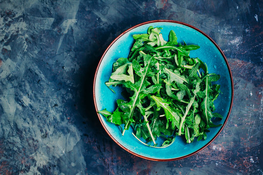Fresh arugula leaves in blue plate. Green vegan salad on a dark stone table background. Top view, copy space