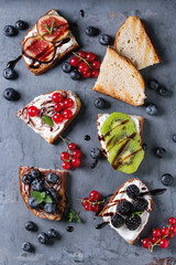 Variety of dessert sandwiches with berries and cream cheese and chocolate swirl. Red currant, blueberries, sliced kiwi, figs over gray metal background. Flat lay, summer appetizer concept