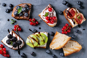 Variety of dessert sandwiches with berries and cream cheese and chocolate swirl. Red currant, blueberries, sliced kiwi, figs over gray metal background. Flat lay, summer appetizer concept