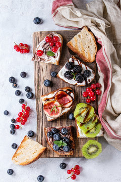 Variety of dessert sandwiches with berries and cream cheese and chocolate swirl. Red currant, blueberries, sliced kiwi, figs on serving board over white texture background. Flat lay, summer appetizer