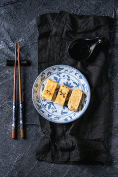 Japanese Rolled Omelette Tamagoyaki sliced with black seasame seeds and soy sauce, served in blue white ornate ceramic plate with chopsticks, textile over black stone background. Top view with space