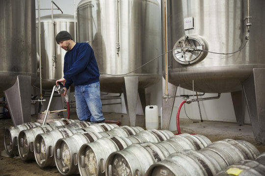A man filling metal beer kegs from large fermentation tanks in a brewery. 