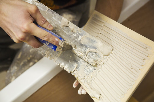 Close up of a builder, tiler sitting in a bathtub spreading adhesive on a tile.