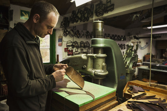 Man standing in a shoemaker's workshop, cutting brown leather.