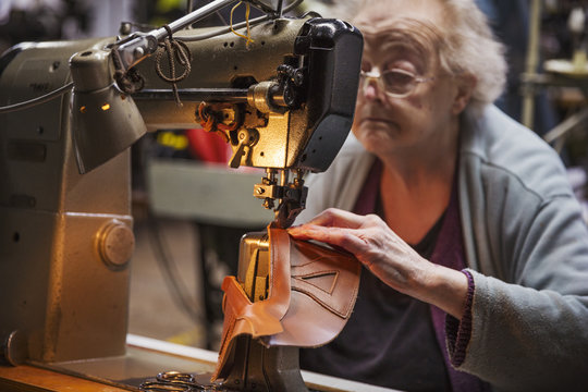 Older woman sitting at a sewing machine in a shoemaker's workshop.