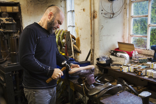Man standing in a shoemaker's workshop by a window, using a hammer on the soles of a work boot.
