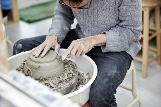 A man using a pottery wheel, shaping a pot base with a small handheld tool shaving off excess clay.  