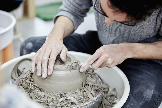 A man seated at a potter's wheel working and shaping a clay pot by removing excess clay. 