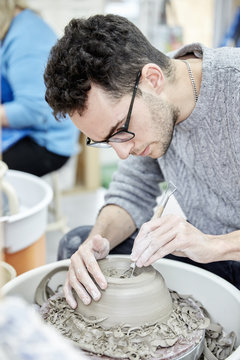 A man seated at a potter's wheel working and shaping a clay pot by removing excess clay. 