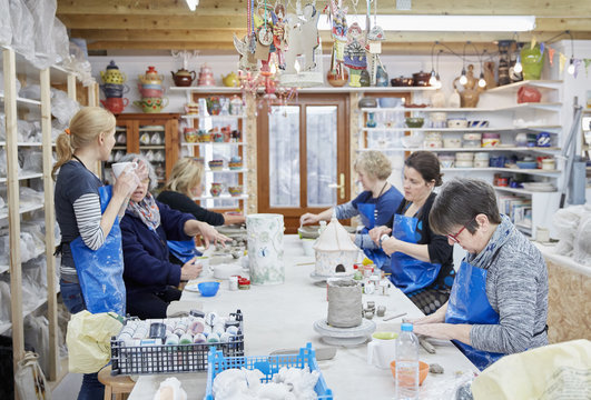 A group of people seated at a workbench in a pottery workshop, handbuilding clay objects. A woman with a cup of tea. 