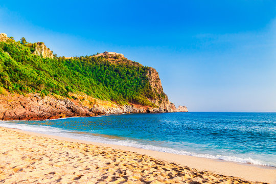 Cleopatra beach on sea coast with green rocks in Alanya peninsula, Antalya district, Turkey. Beautiful sunny landscape for tourism with clear water and sand. Alanya Castle on the cliff