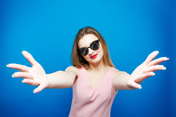 Cute Brunette Wearing Pink Dress, Sunglasses and Sport Shoes is Showing Stop by Hands on Blue Background in Studio.