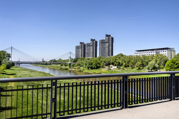 Fototapeta na wymiar Germany, Baden-Wuerttemberg, Mannheim, Neckarsteg (Collini-Steg): Panoramic view from bridge with Neckar river, metal fence, skyscrapers, green park and blue sky