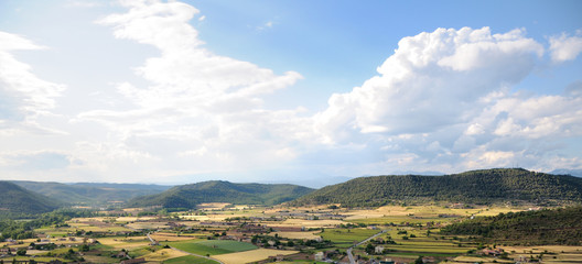Obraz premium Sunlit large valley with colorful fields and small houses. The valley is surrounded by green mountains. Blue sky and white clouds. Summer. Spain.