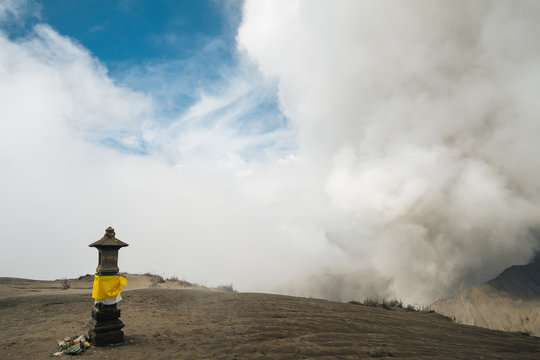 sacred stone pole on the top of Batok and background view of Bromo erupting and bright blue sky. Ashes flow above the volcano, covering blue sky. Sand on the ground and yellow fabric on the altar