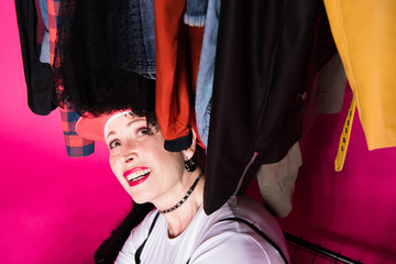 happy senior woman sitting under clothes on hangers and looking on them isolated on pink