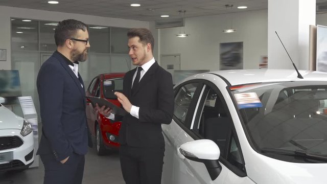 Attractive sales manager explaining the contract to the customer at the dealership. Handsome male seller dresed in black suit telling the client about the equipment of the car. Young caucasian