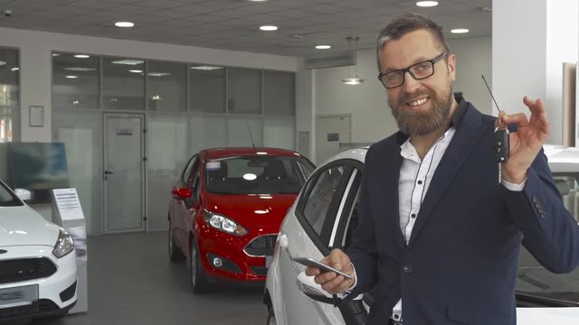 Attractive bearded man calling on the phone at the car dealeship. Handsome male customer showing key of the vehicle. Mature guy in glasses and stylish suit standing near the white hatchback