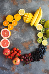Rainbow colored fruits in a circle, strawberries, blueberries, mango, orange, grapefruit, banana, apple, grapes, kiwis, papaya on the grey background, copy space for text, selective focus