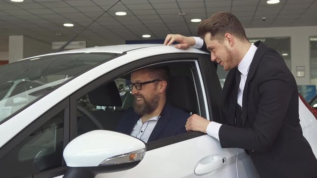 Attractive sales manager consulting the customer about the car at the dealership. Handsome male seller in black suit standing near the vehicle while the client sitting inside it. Mature caucasian man