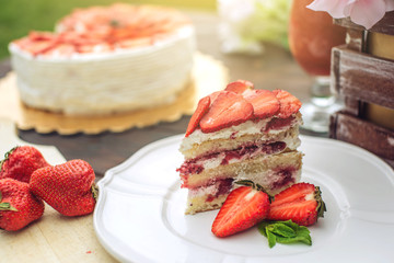 Delicious homemade summer cake with strawberries and butter cream on a wooden porch and a piece on the plate