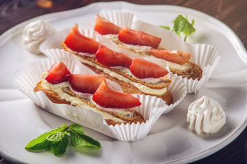 Delicious homemade cakes eclairs with strawberries and cream on a wooden porch and a piece on the plate