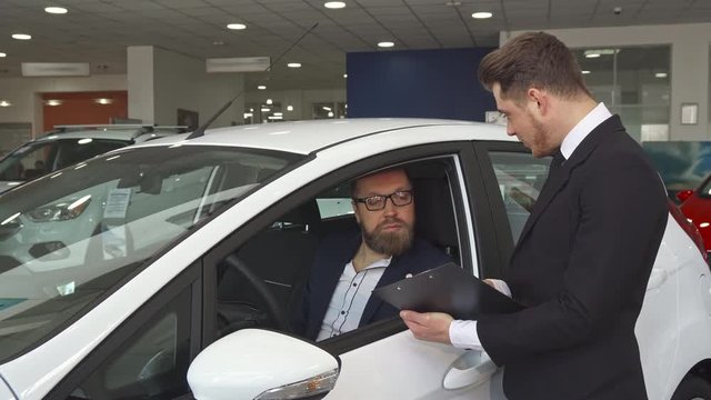 Handsome sales manager showing customer something on clipboard. Attractive bearded man sitting inside the white hatchback at the showroom. Young male seller in black suit instructing his client about