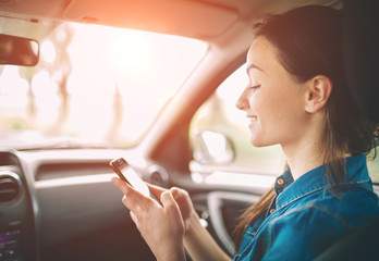 Beautiful woman smiling while sitting on the front passenger seats in the car. Girl is using a smartphone - obrazy, fototapety, plakaty