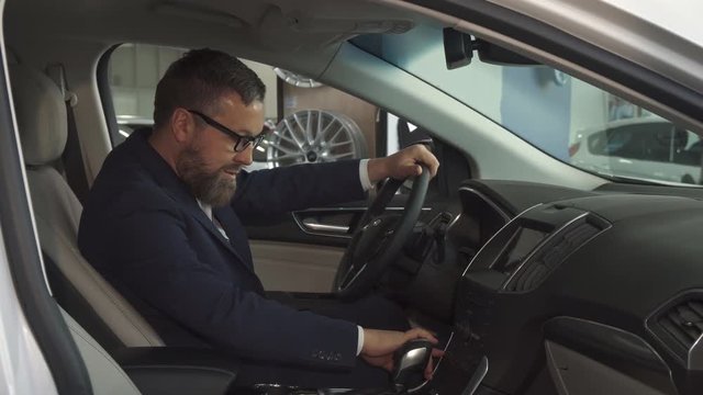 Male bearded customer approving car interior at the dealership. Attractive caucasian man in glasses showing his thumb up inside th vehicle. Handsome brunette guy pushing some buttons on the console