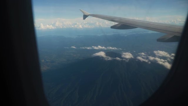 View through an airplane window on the crater volcano, mountains, sky and clouds. Rocky Mountains through Airplane Window. Aerial view Clouds and sky as seen through window of an aircraft. 4K video