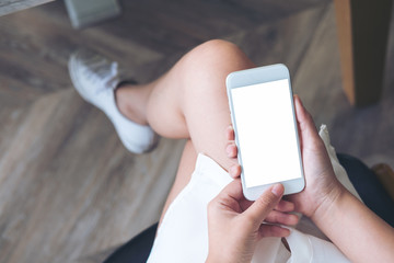 Mockup image of woman's hand holding white mobile phone with blank screen on thigh with wooden floor background in modern