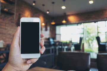 Mockup image of hand holding white mobile phone with blank black screen in modern cafe
