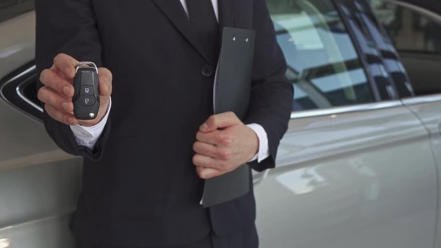 Close up of sales manager showing key near the new car. Low shot of male seller holding clipboard near his body. Man dressed in black suit and tie standing near the gray sedan